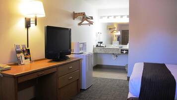 A hotel room with a bed, a desk with a television, a microwave, a small refrigerator, a mirror, and a hanging coat rack.