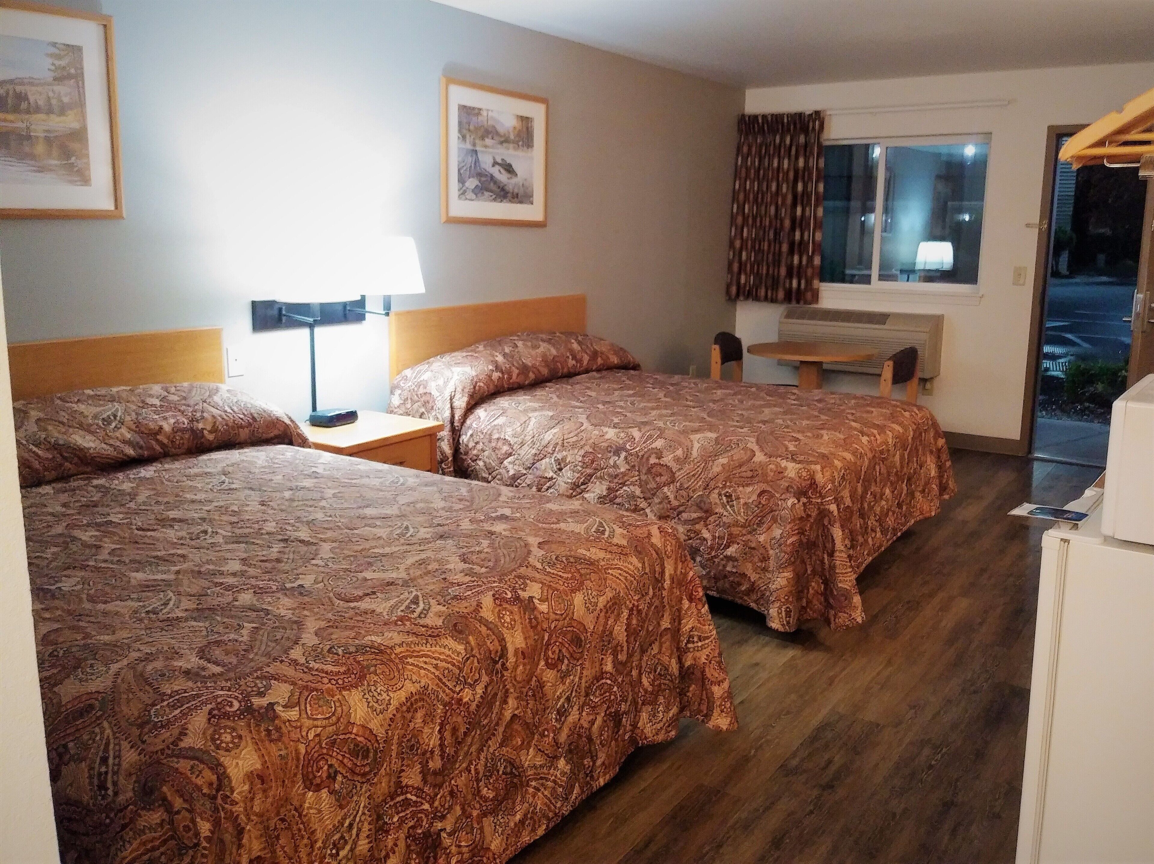 Une chambre d’hôtel avec deux lits, un bureau, une chaise, une fenêtre avec des rideaux et une porte donnant sur l’extérieur.
