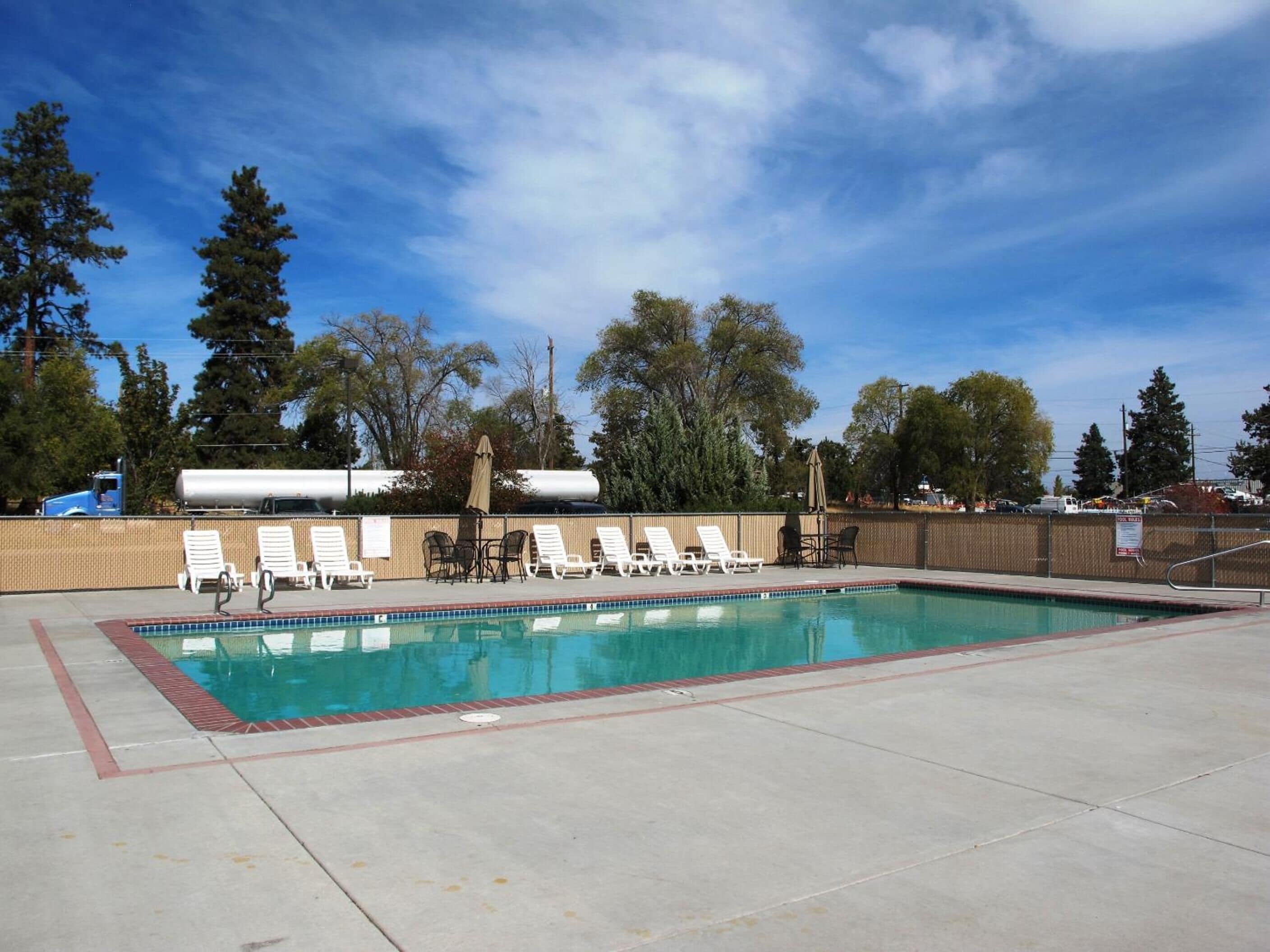 Una piscina rectangular al aire libre con borde de azulejos, rodeada por un muro de ladrillo bajo. Hay varias sillas reclinables blancas y una mesa con sillas, algunas con sombrillas. El área está delimitada por una valla, y se ven árboles y un cielo despejado al fondo.