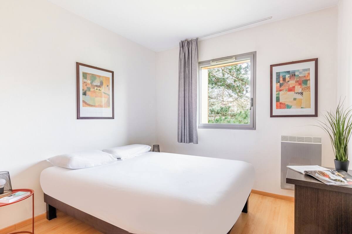 A modern hotel room with a large bed, a desk with a plant, and two framed artworks on the walls.