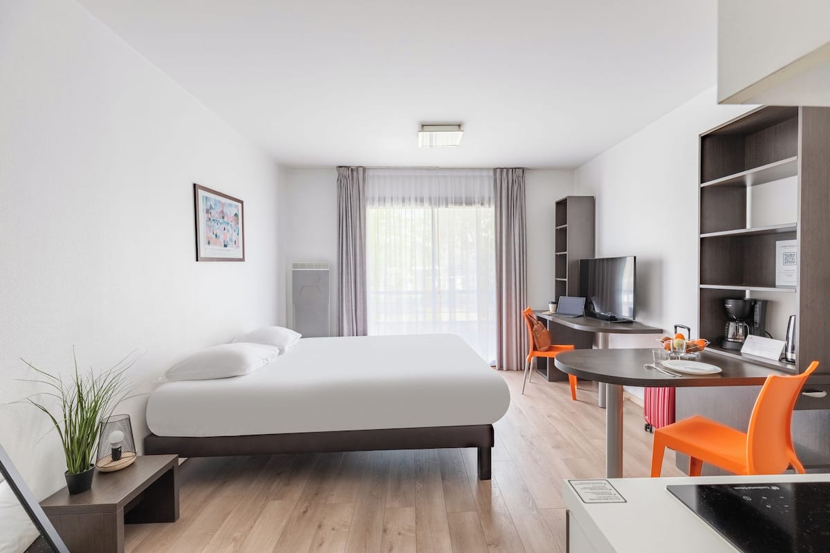 A modern hotel room with a large bed, a desk with a chair, a television, and a bookshelf.