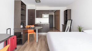 A compact hotel room with a kitchenette, a bed, a desk with a chair, and a suitcase.