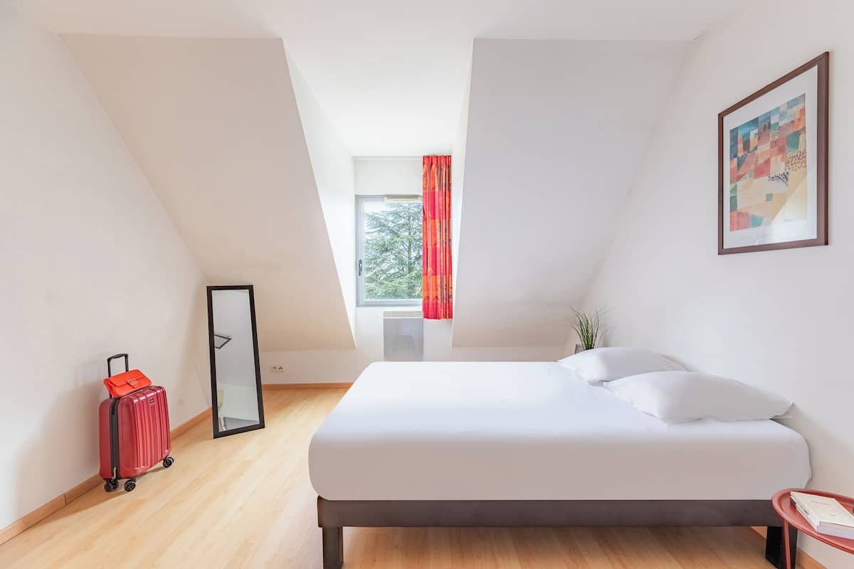 A modern hotel room with a bed, a red suitcase, a window with curtains, and a framed picture on the wall.