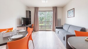 A modern living room with a grey sofa, orange chairs, a dining table set for two, a TV, and a large window with curtains.