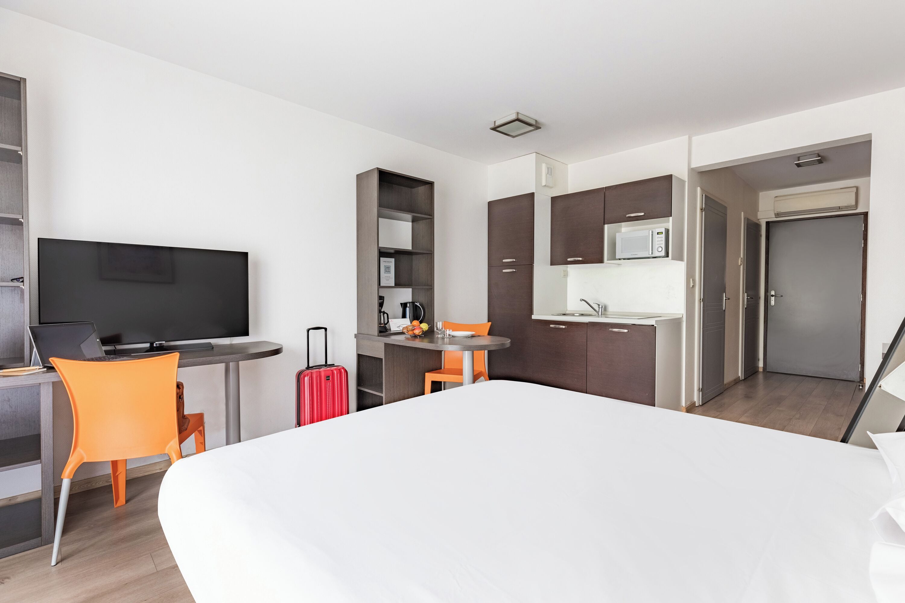 A hotel room with a bed, a desk with a laptop, a TV, a small kitchen area with a microwave, and a suitcase.