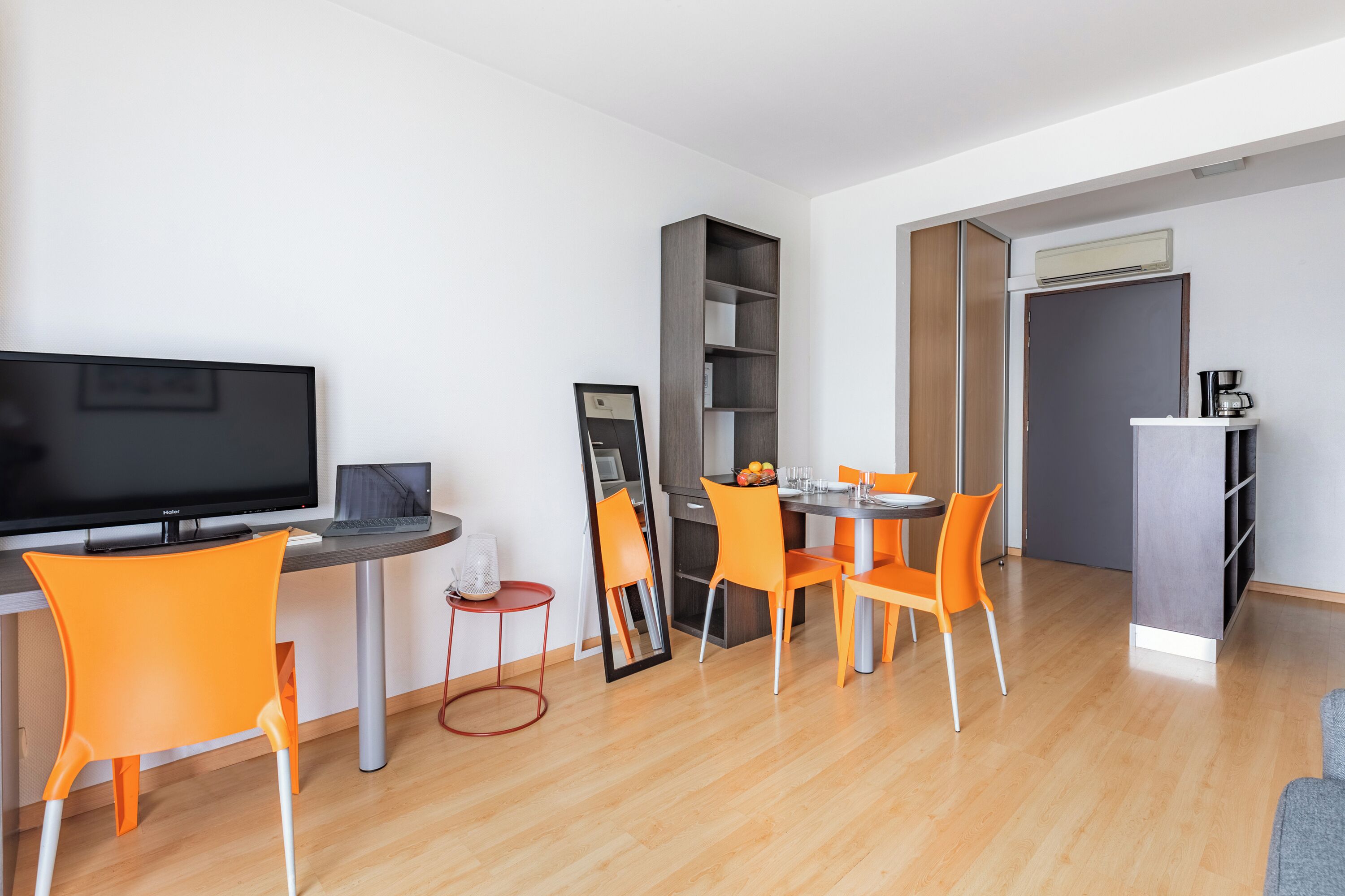 Een moderne woonkamer met een flatscreen televisie, een eettafel met oranje stoelen, een boekenkast en een keukenhoek met een koffiezetapparaat.