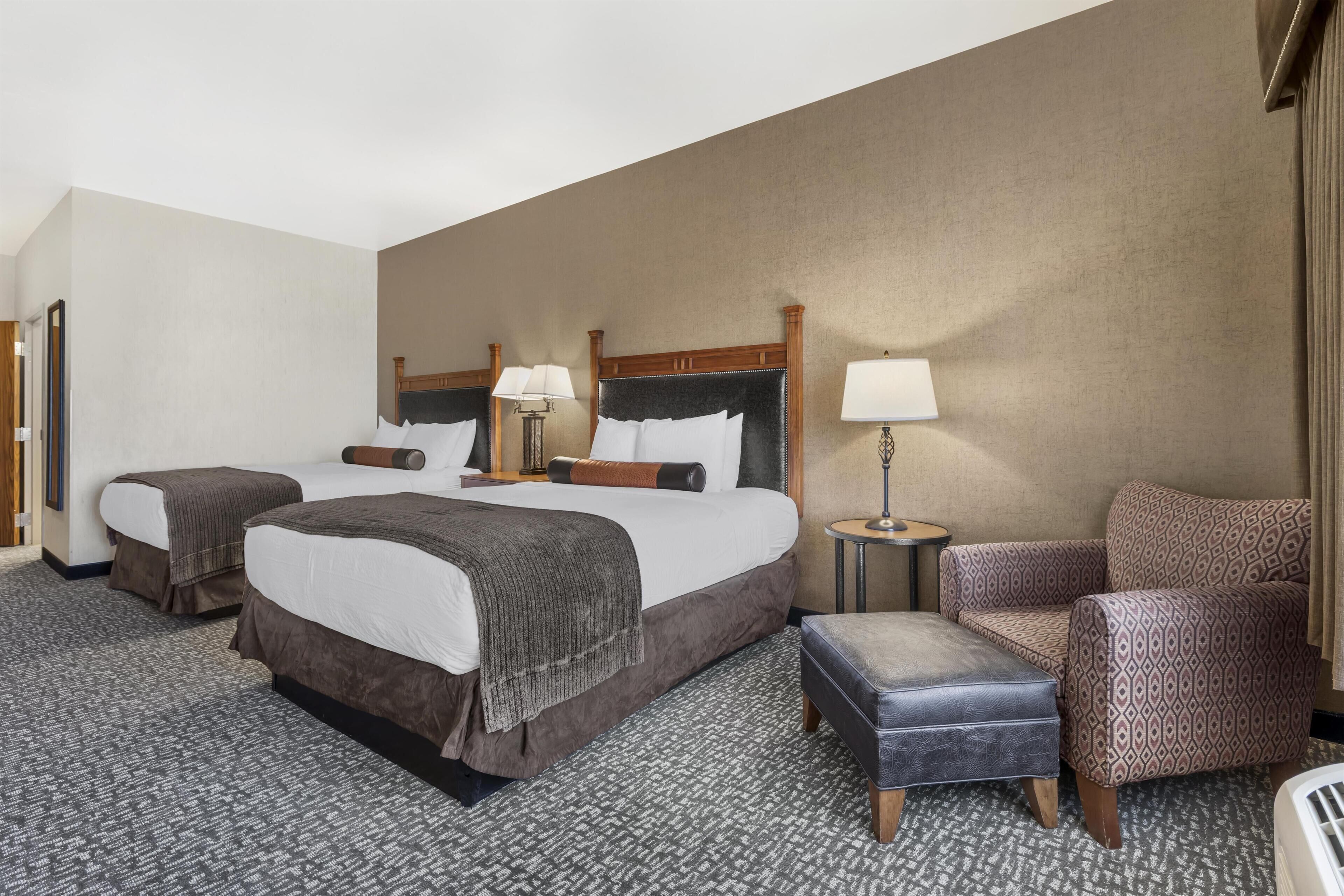 A hotel room with two beds, a chair, a small table, and a lamp.