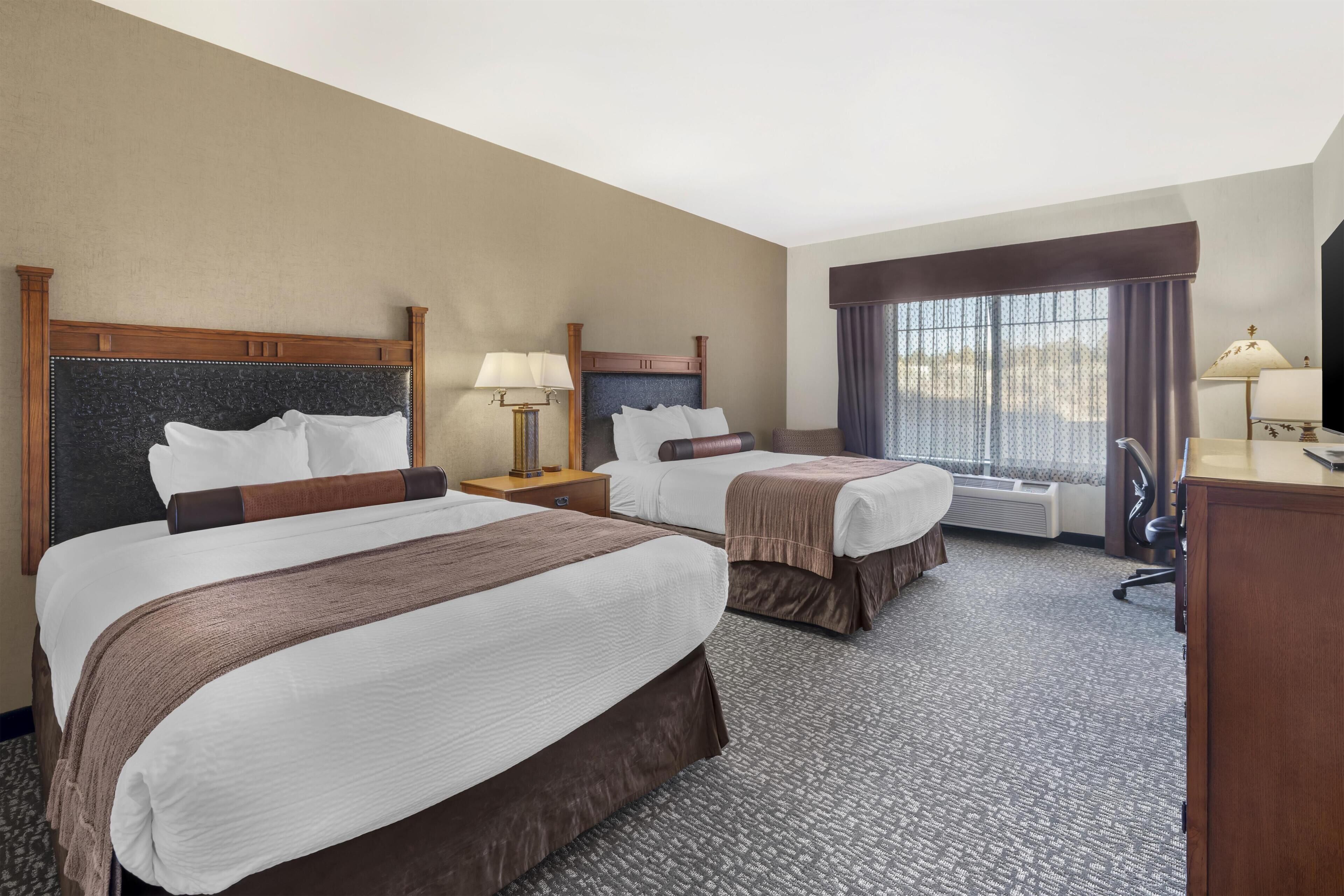 A hotel room with two beds, a desk, a chair, a lamp, a window with curtains, and a heater.