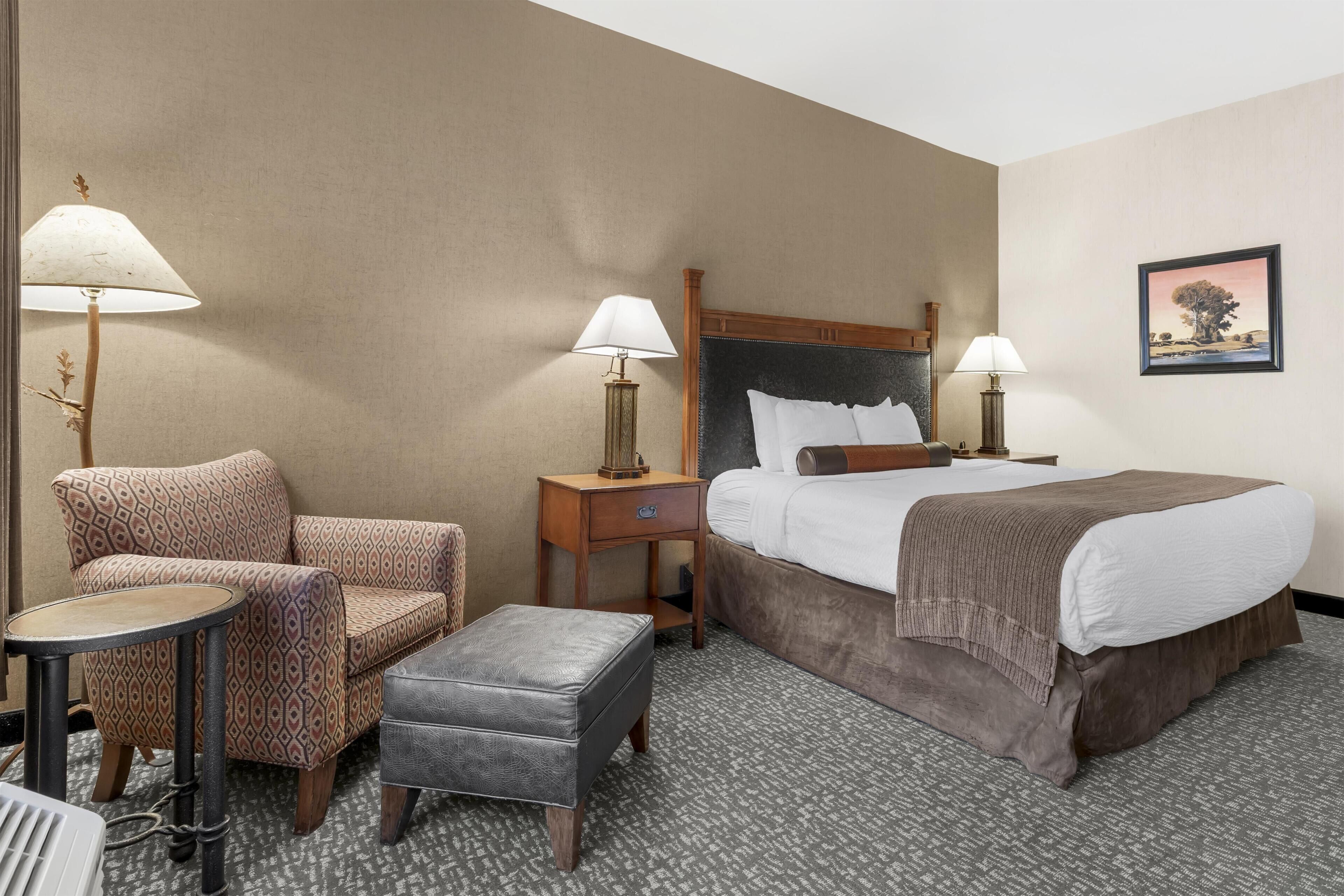 A hotel room with a bed, two bedside tables with lamps, a chair, a small table, and an ottoman.