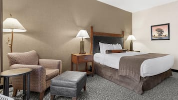 Quarto de hotel com uma cama, duas mesas de cabeceira com abajures, uma cadeira, uma mesinha e um banquinho.