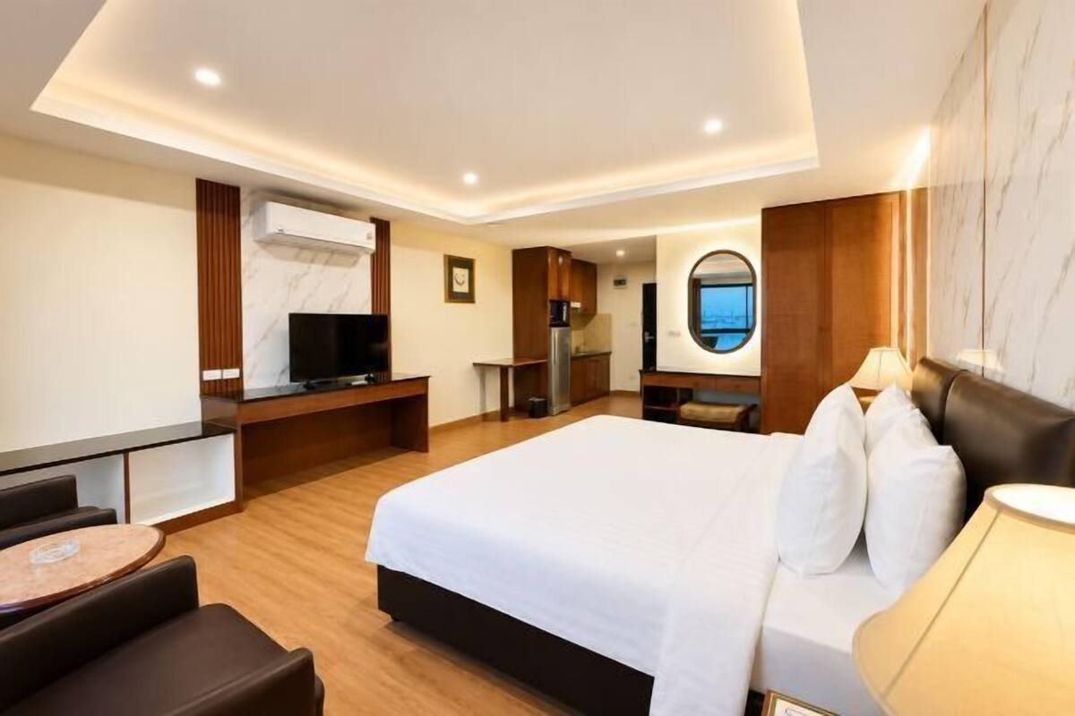 A hotel room with a large bed, a desk, a television, a small kitchen area, and a view of the sky through a window.
