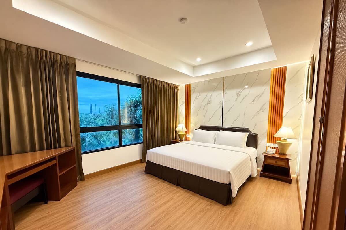 A hotel room with a bed, bedside tables, a desk, a window with curtains, and a view of palm trees and buildings.