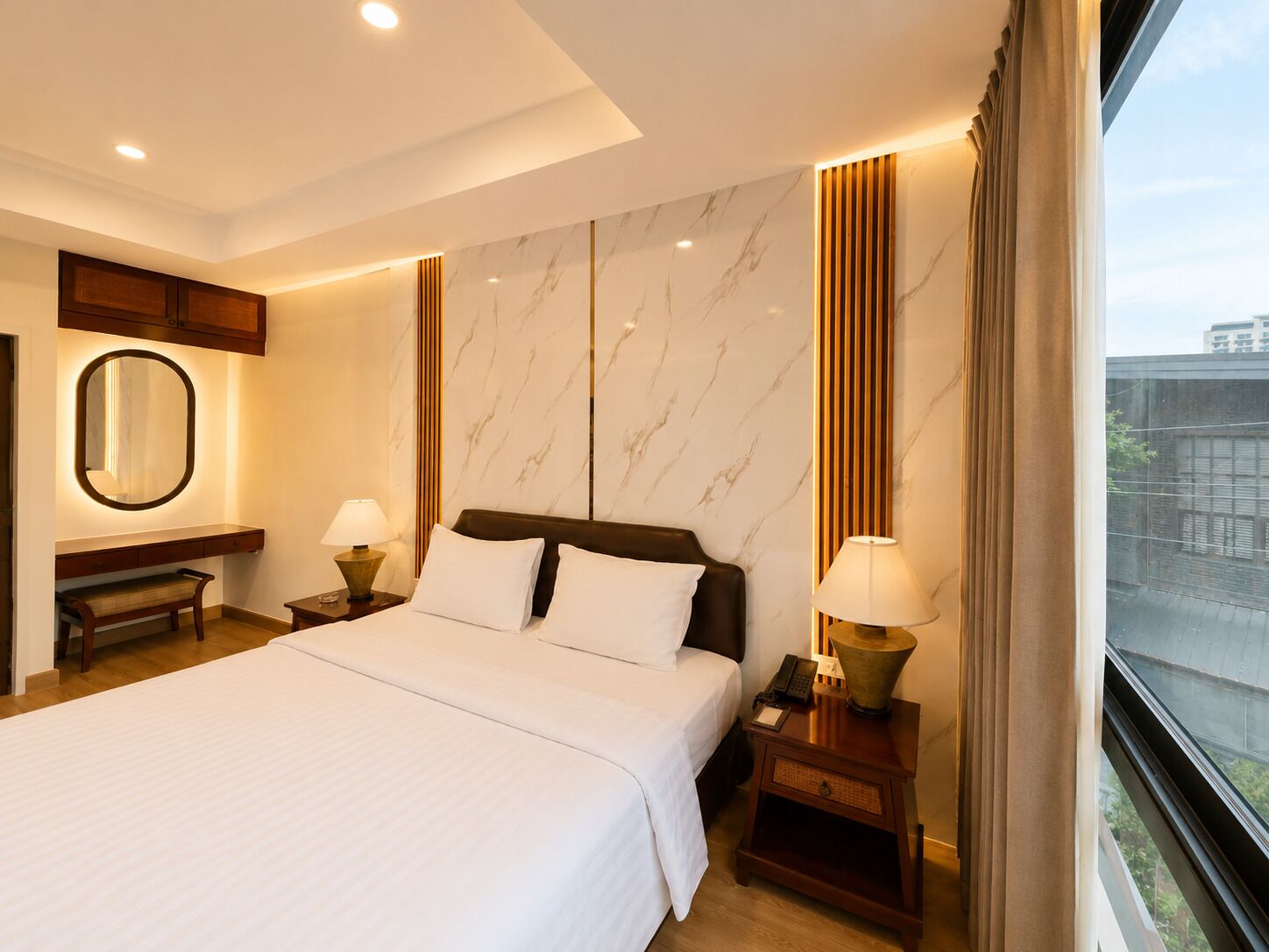 A hotel room with a large bed, bedside tables with lamps, a mirror, and a view of the city.
