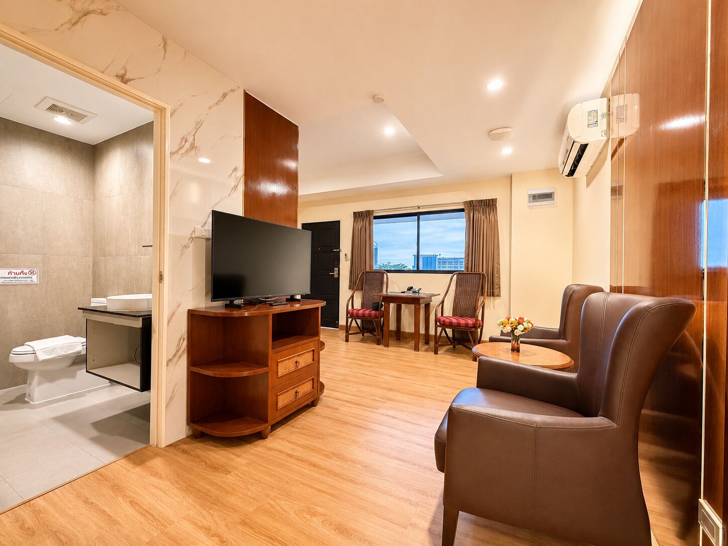 A hotel room with a wooden entertainment unit, a television, a dining area with a table and chairs, and a large window with a view.
