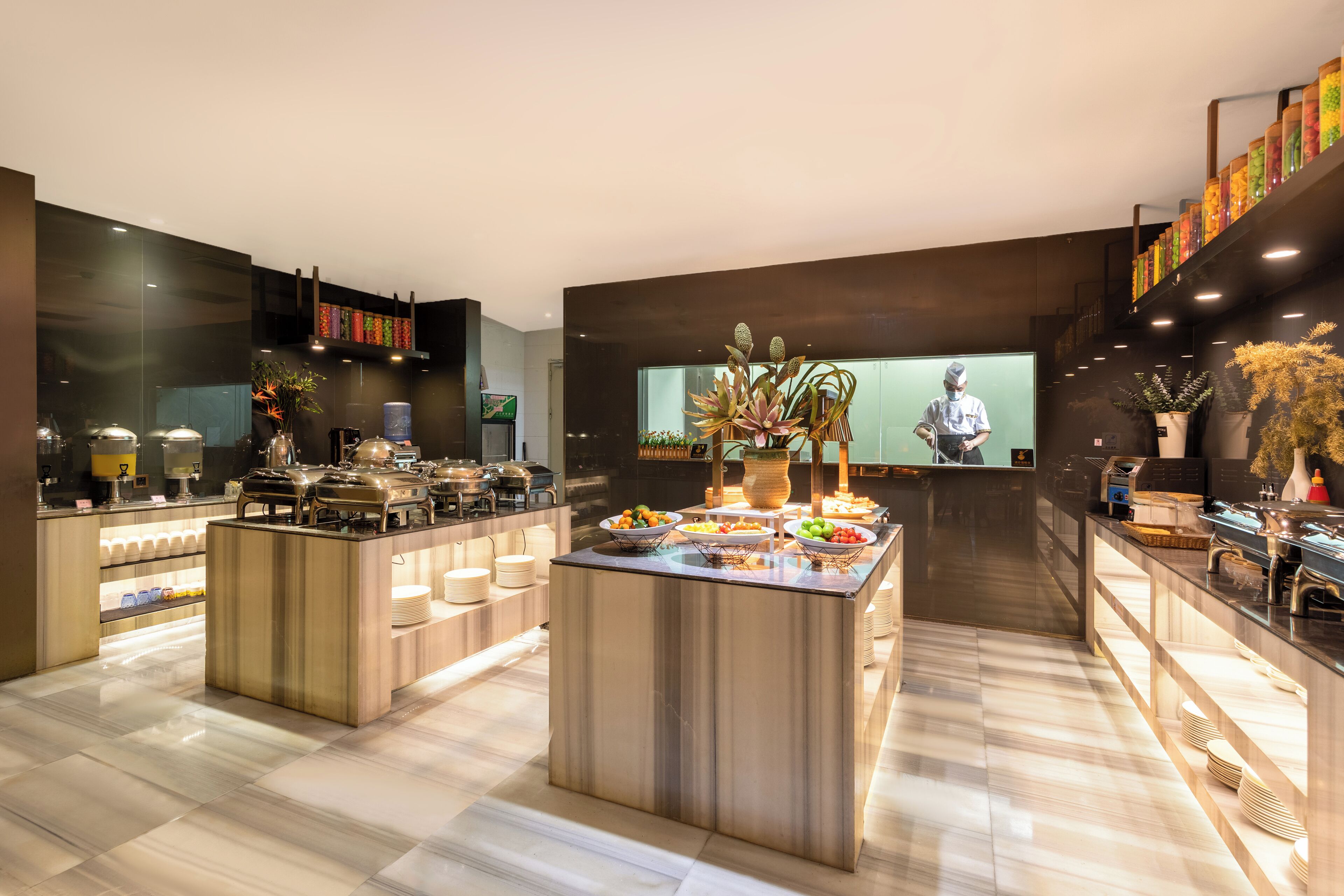 A modern hotel lobby with a central buffet area, a reception desk, and a staircase with colorful decorations.