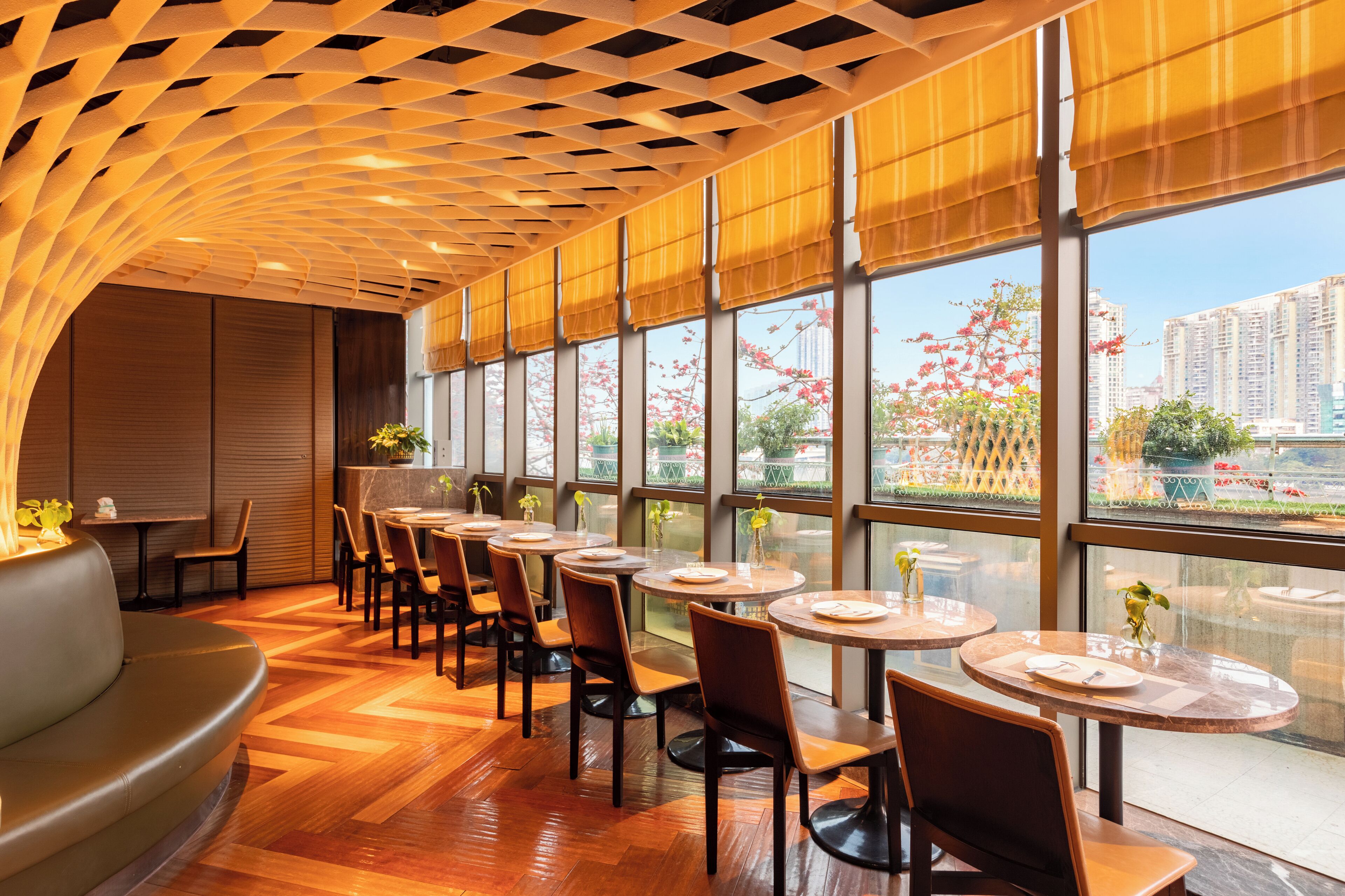 A restaurant with round tables and chairs, large windows with a view of buildings and trees, and a wooden ceiling.