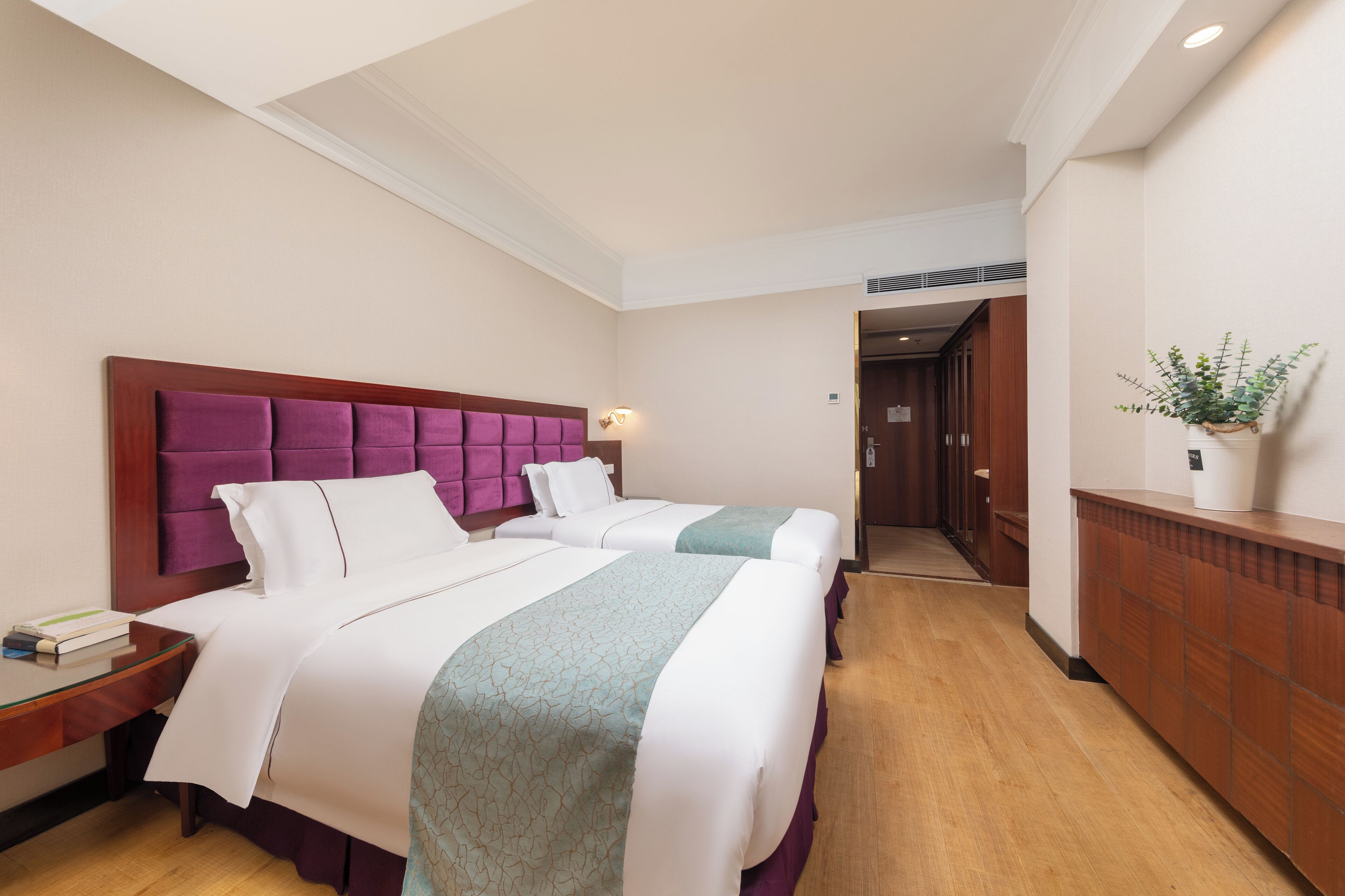 A hotel room with two beds, a wooden headboard, a bedside table, a nightstand, and a potted plant on a wooden dresser.