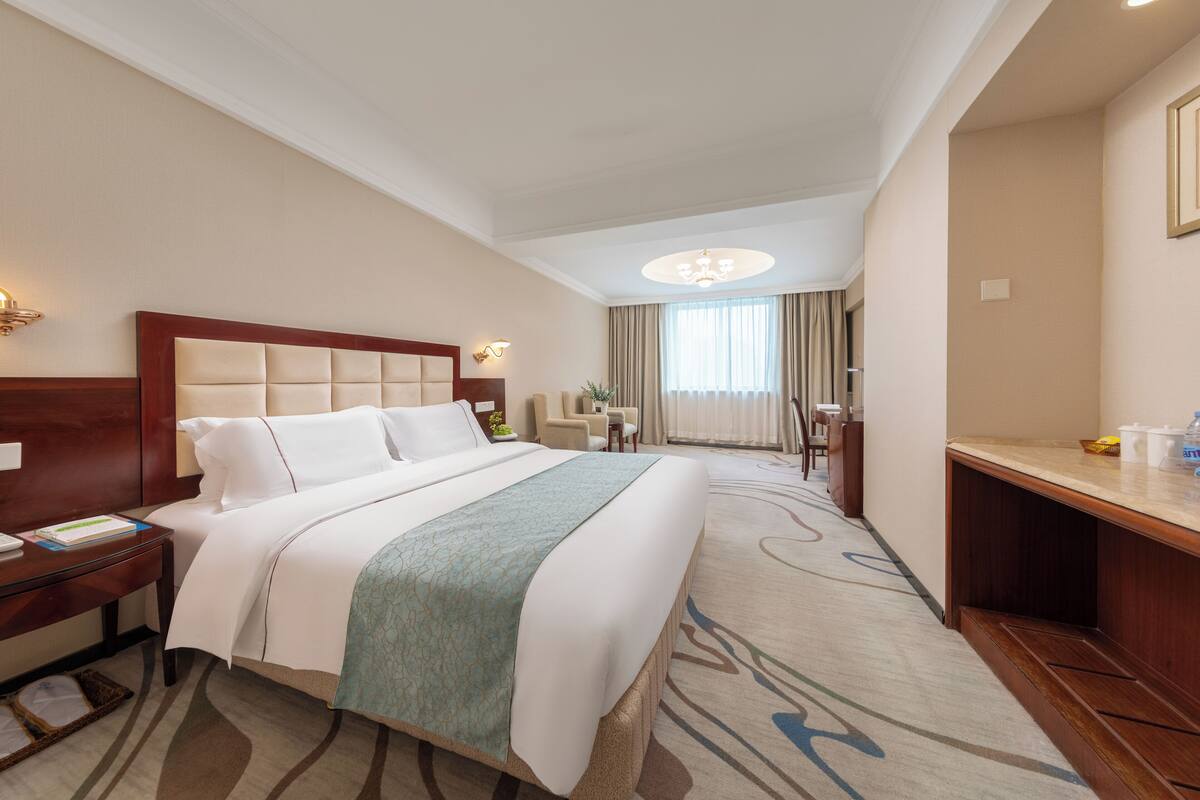 A hotel room with a large bed, a desk, a chair, and a small table. There is a window with curtains and a chandelier on the ceiling.