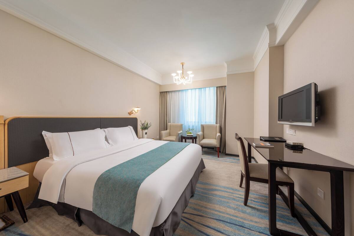 A hotel room with a large bed, a desk, a chair, a television, and a small table with a plant.