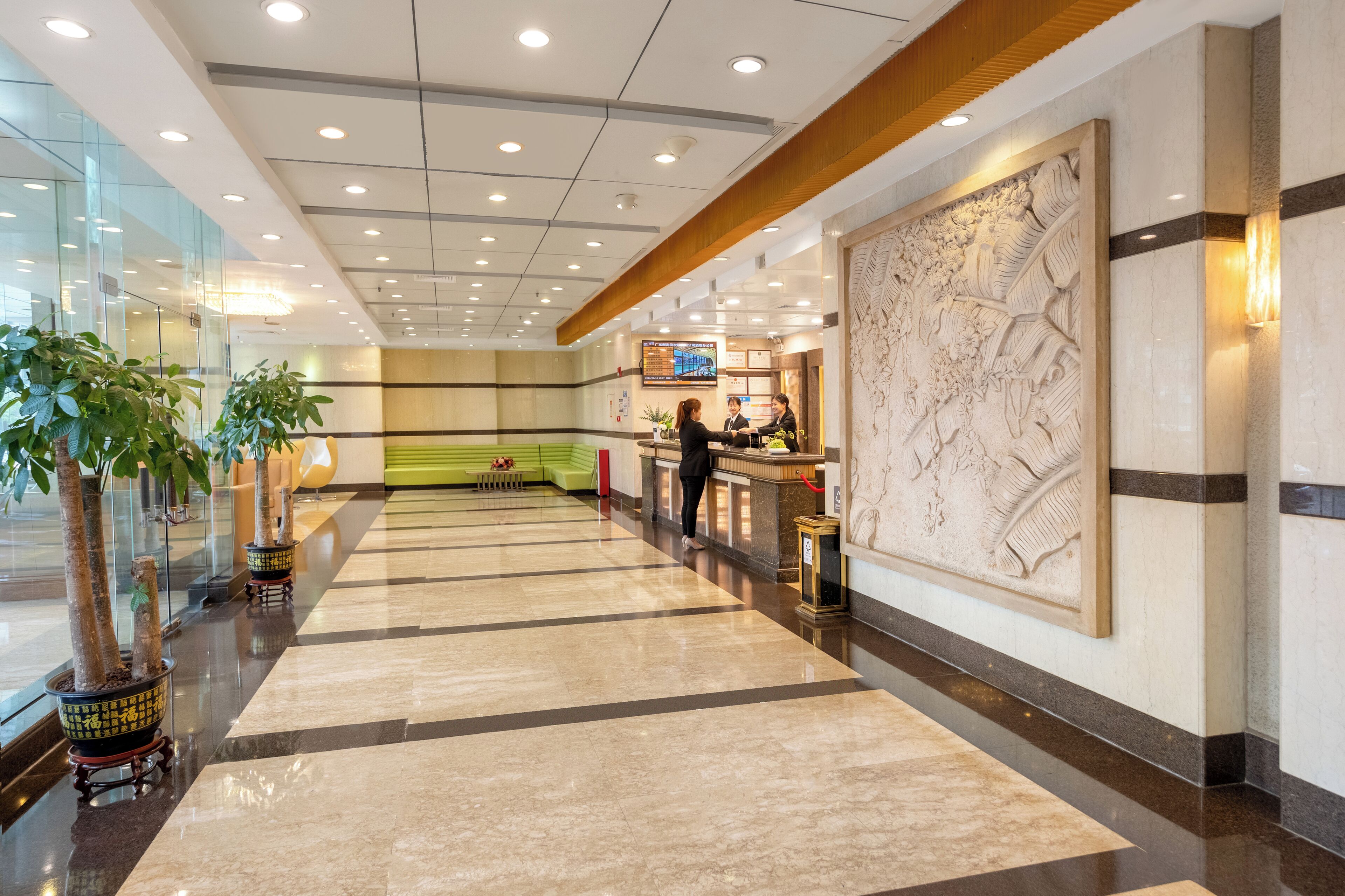 A hotel lobby with a reception desk, potted plants, a large decorative wall, and a polished floor.