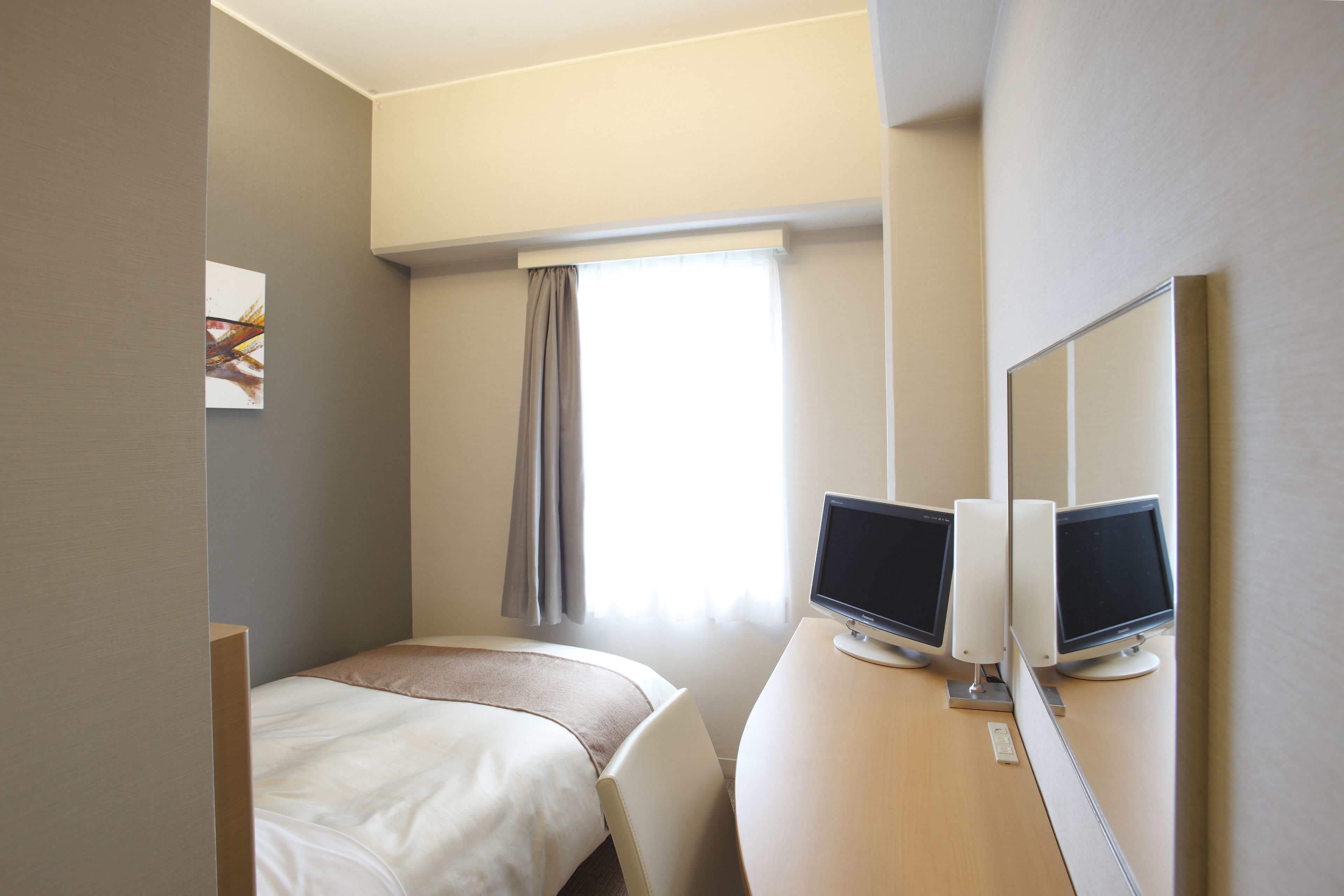 A hotel room with a bed, a desk with two monitors, a telephone, and a window with curtains.