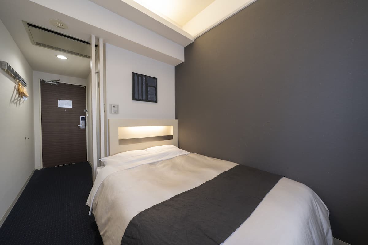Une chambre d’hôtel avec un lit, une table de chevet, une commode, une lampe fixée au mur, une porte et une petite fenêtre.