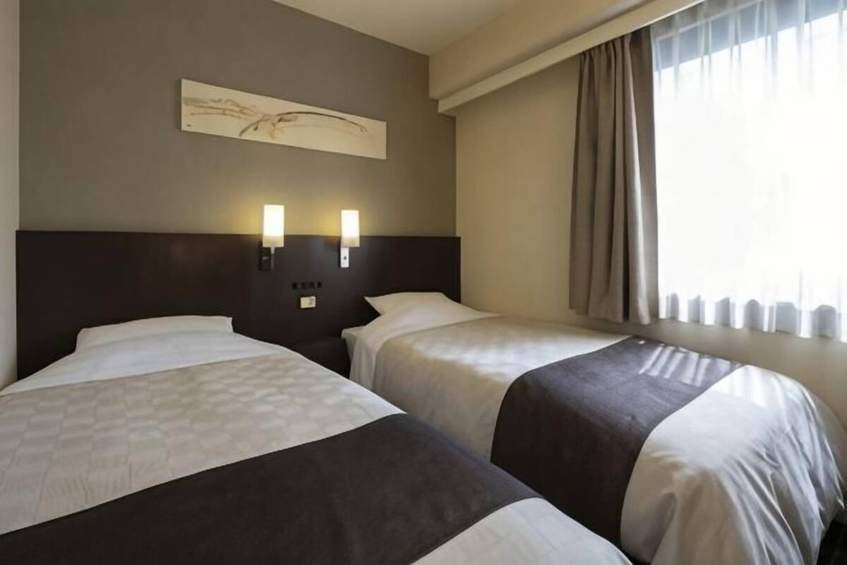 Une chambre d’hôtel avec deux lits, une tête de lit, des lampes fixées au mur, une fenêtre avec des rideaux et un tableau accroché au mur.