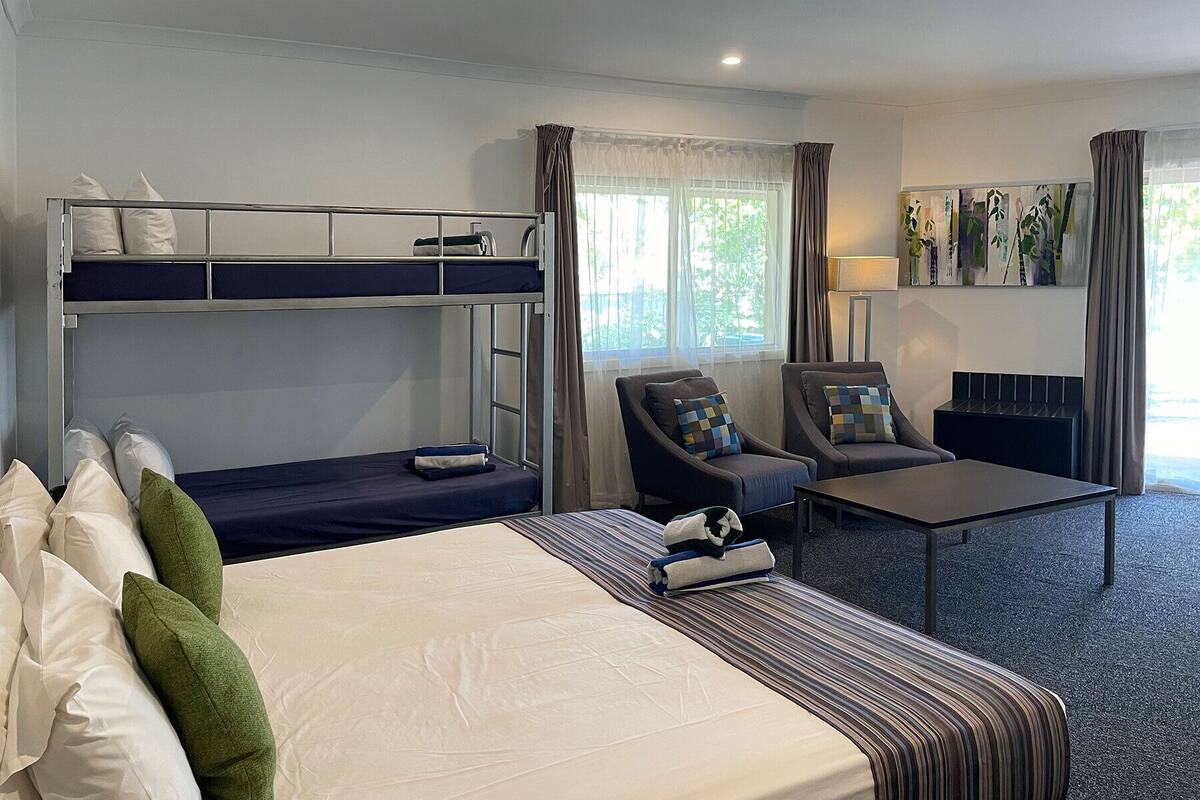 Una habitación de hotel con una cama litera, una cama individual, una silla, una mesita pequeña y una lámpara.
