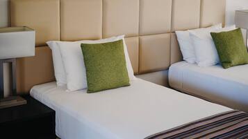 Una habitación de hotel con dos camas, cada una con ropa blanca y almohadas verdes, un cabecero color beige y una mesita de noche con una lámpara.