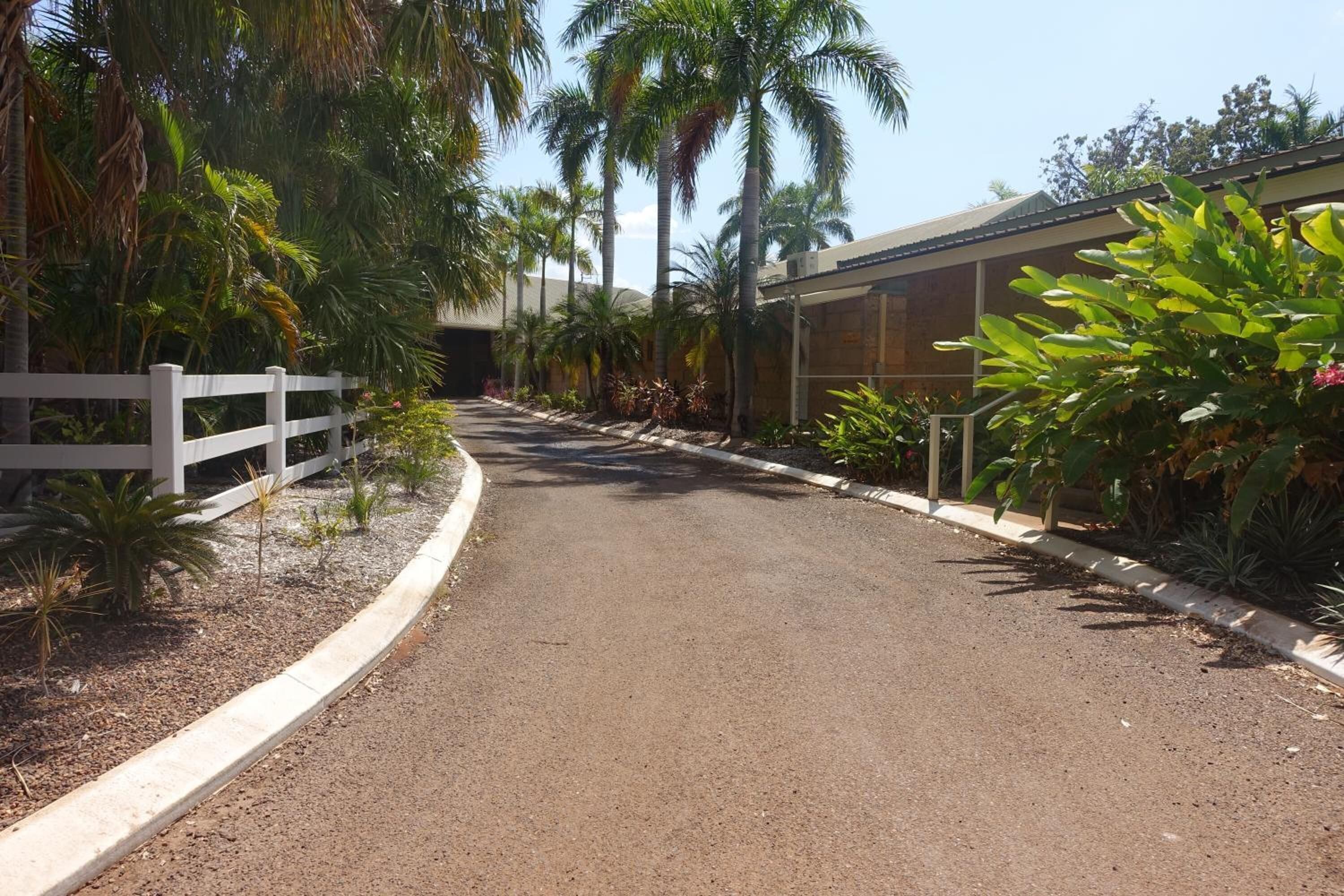 Un chemin pavé bordé d’une clôture blanche et de diverses plantes tropicales, qui mène à un bâtiment doté d’un toit en métal et de fenêtres en verre.