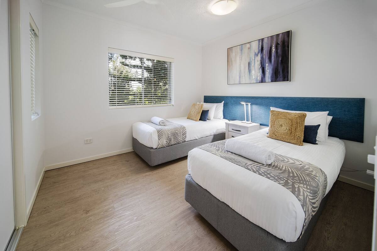A bedroom with two beds, a large window with blinds, a ceiling fan, and a wall-mounted abstract painting.