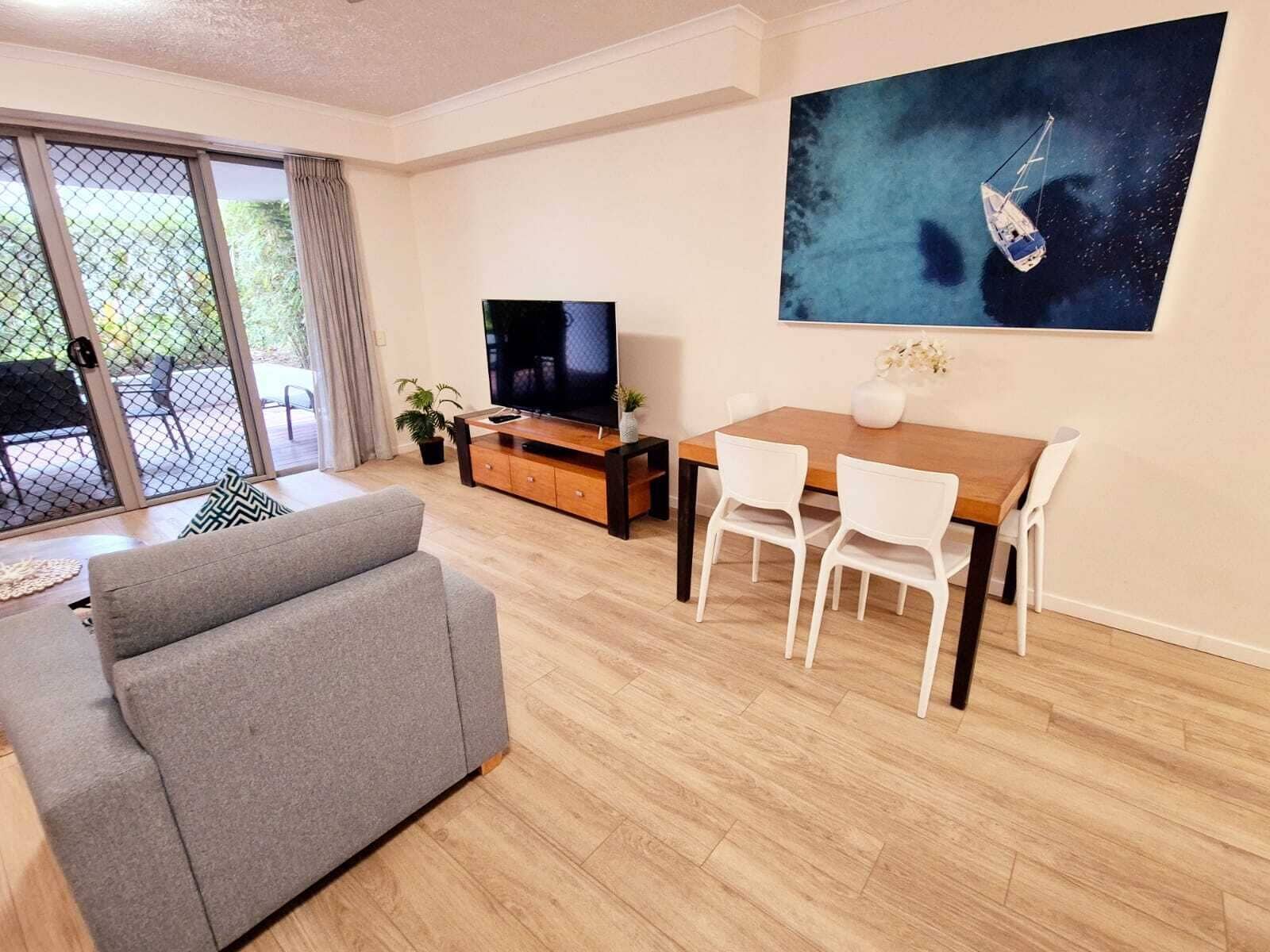 Ein modernes Wohnzimmer mit einem grauen Sofa, einem Holzesstisch mit weißen Stühlen, einem Fernseher auf einem Holzboden und einem großen Gemälde eines Segelbootes an der Wand.