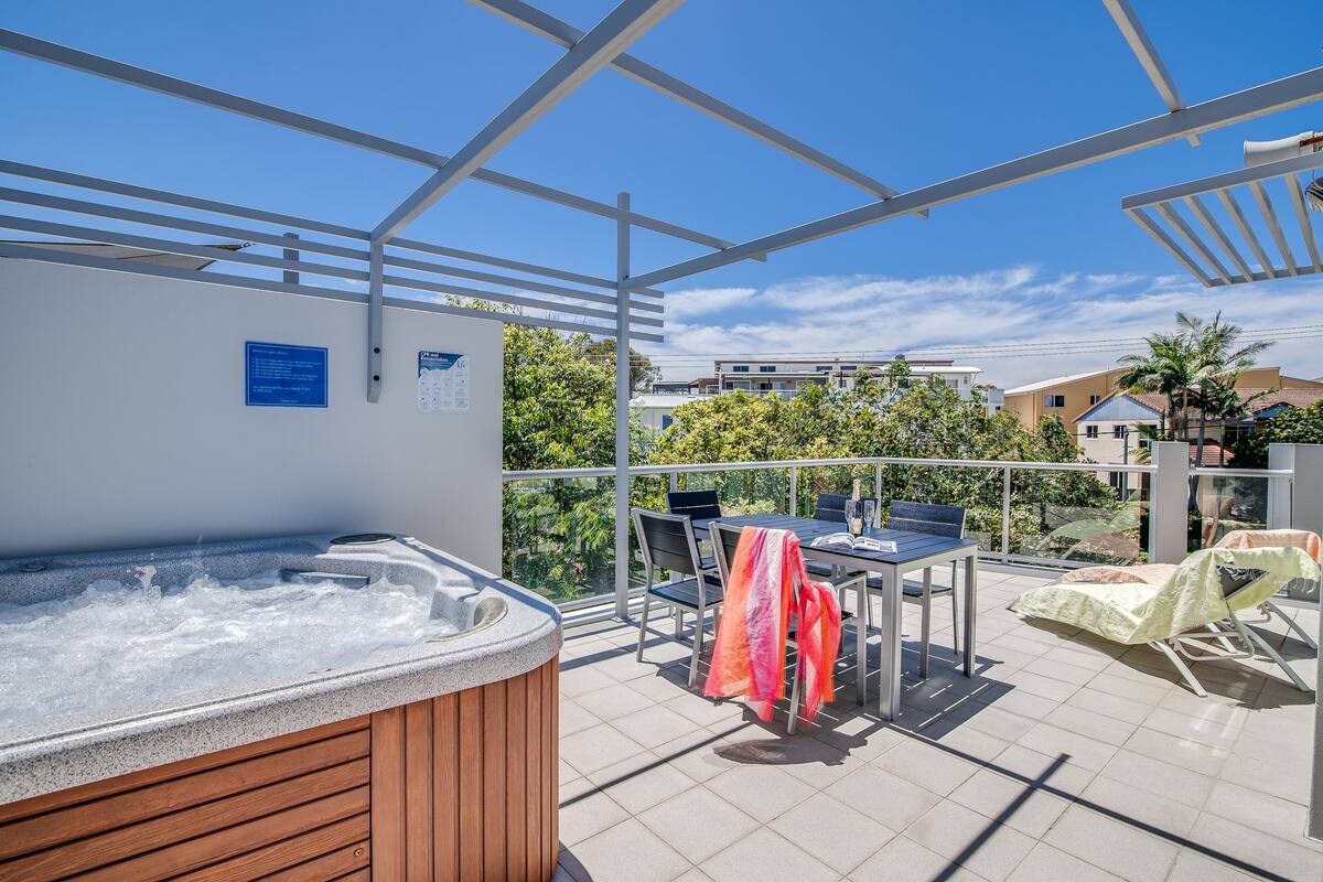A rooftop terrace with a hot tub, outdoor seating, and a view of surrounding buildings and trees.