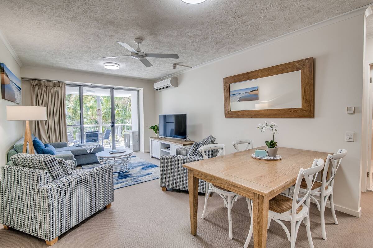 A modern living room with a wooden dining table, white chairs, a sofa, and a television.
