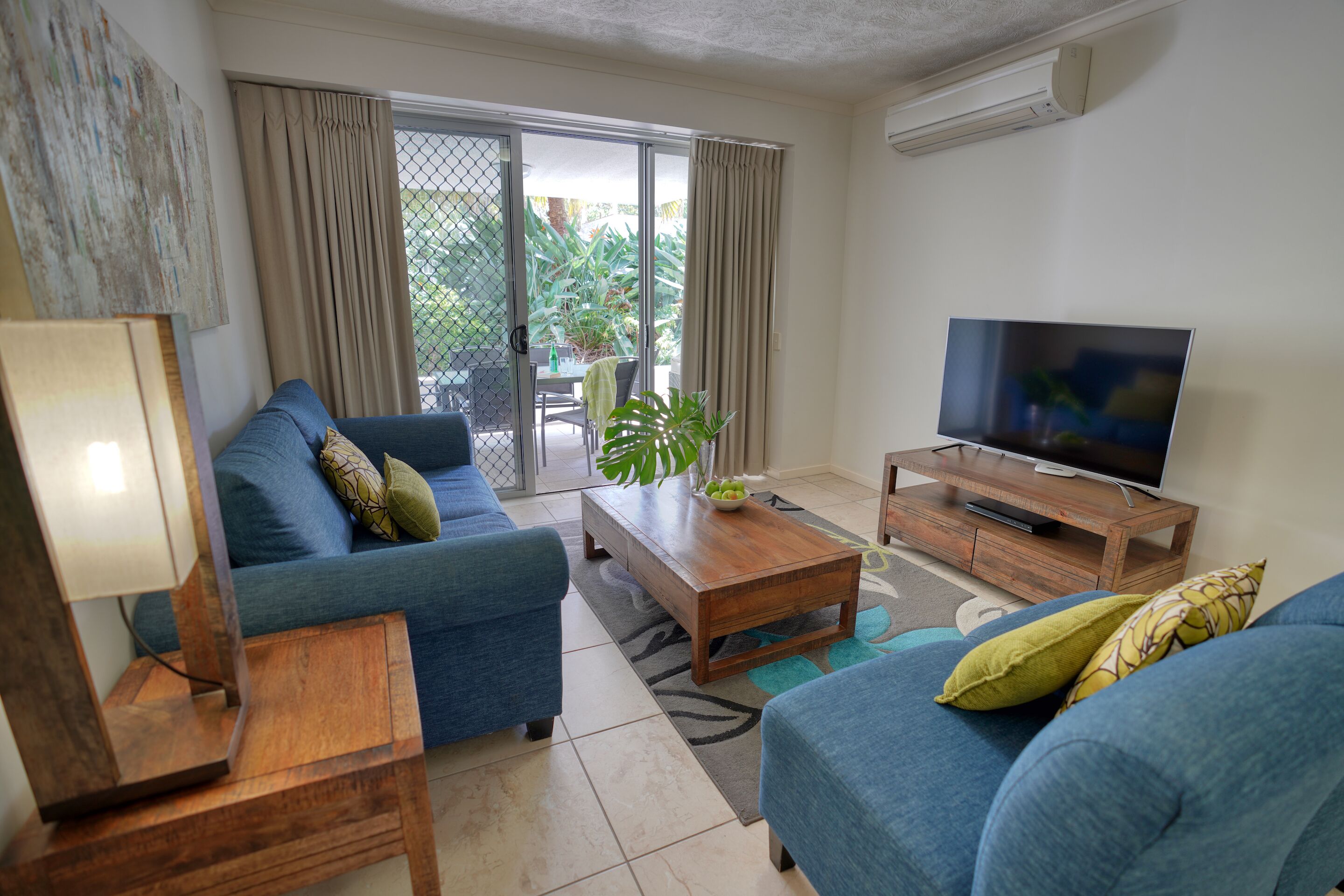 A living room with a blue sofa, a wooden coffee table, a TV, and a sliding glass door leading to a balcony with plants.