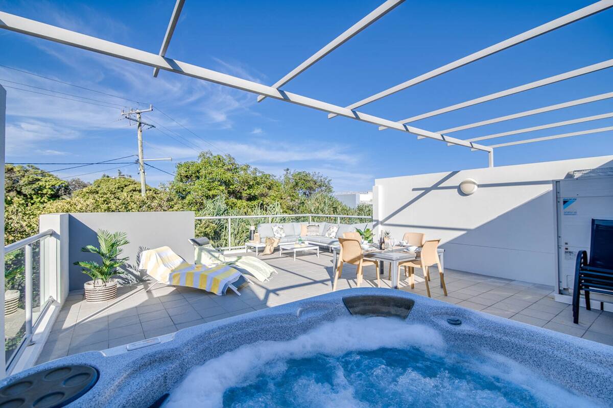 A rooftop terrace with a hot tub, outdoor seating, and a view of the sky and greenery.
