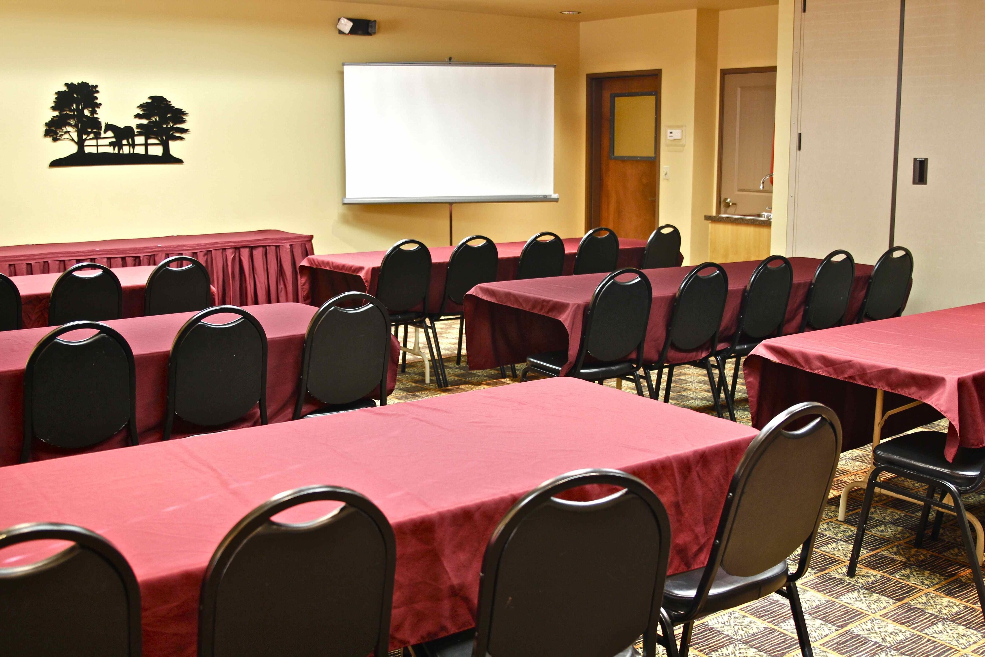Un salón de conferencias con varias mesas y sillas, una pizarra y una pantalla de proyector.