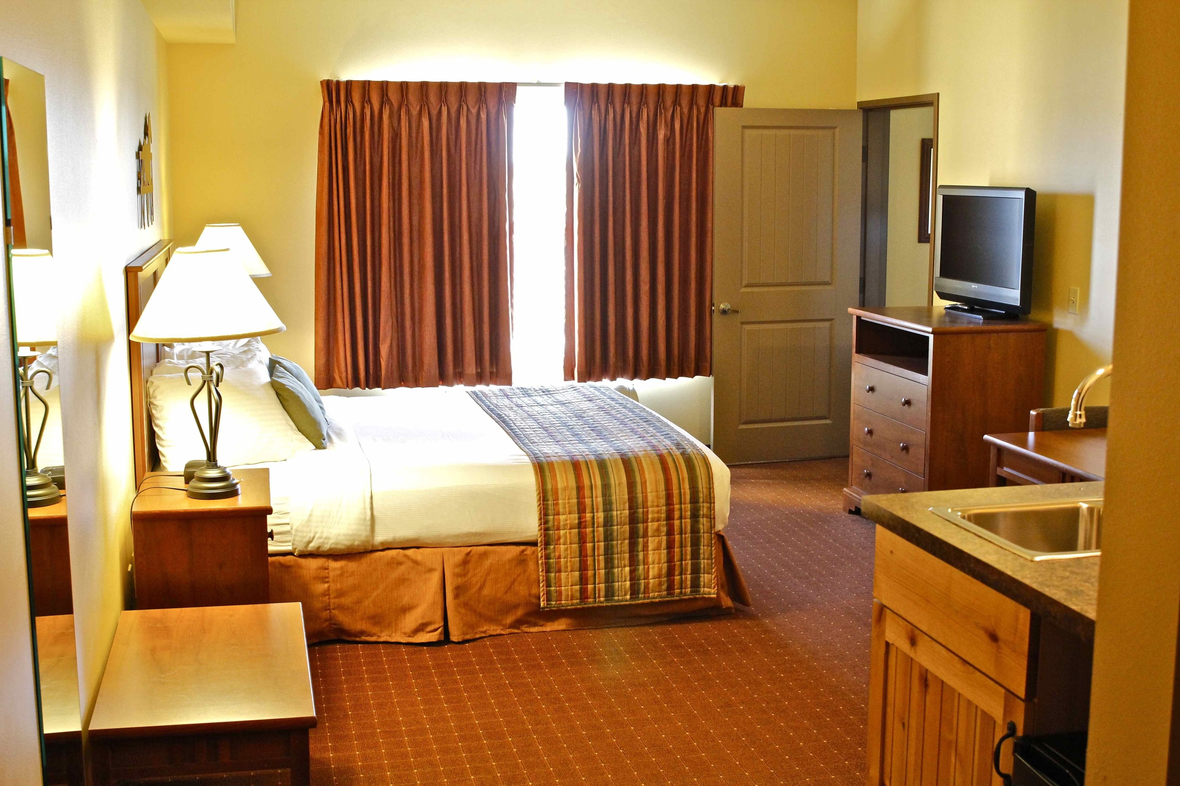 A hotel room with a bed, bedside lamps, a television, a small kitchenette, and a door.