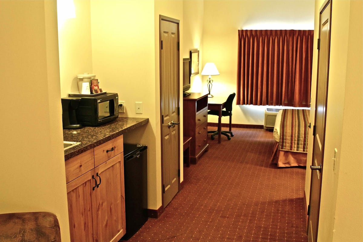 A hotel room with a kitchenette, a desk with a chair, a bed, and a window with curtains.