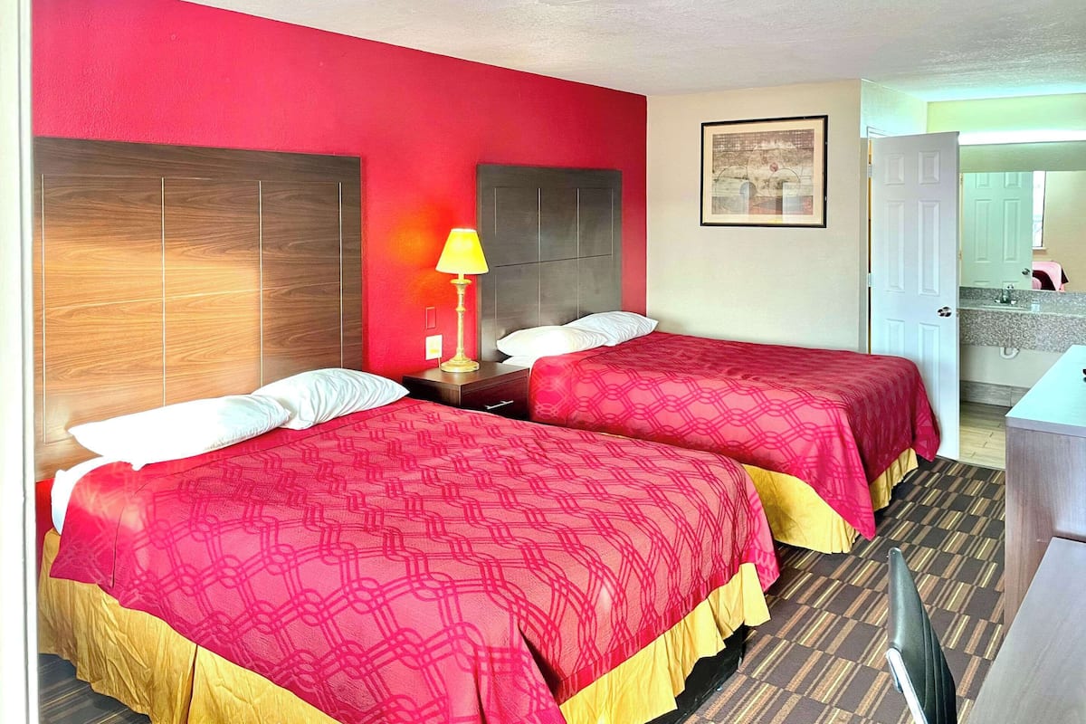 Une chambre d’hôtel avec deux lits, une table de chevet avec une lampe, un tableau encadré au mur et une salle de bain visible à travers une porte ouverte.