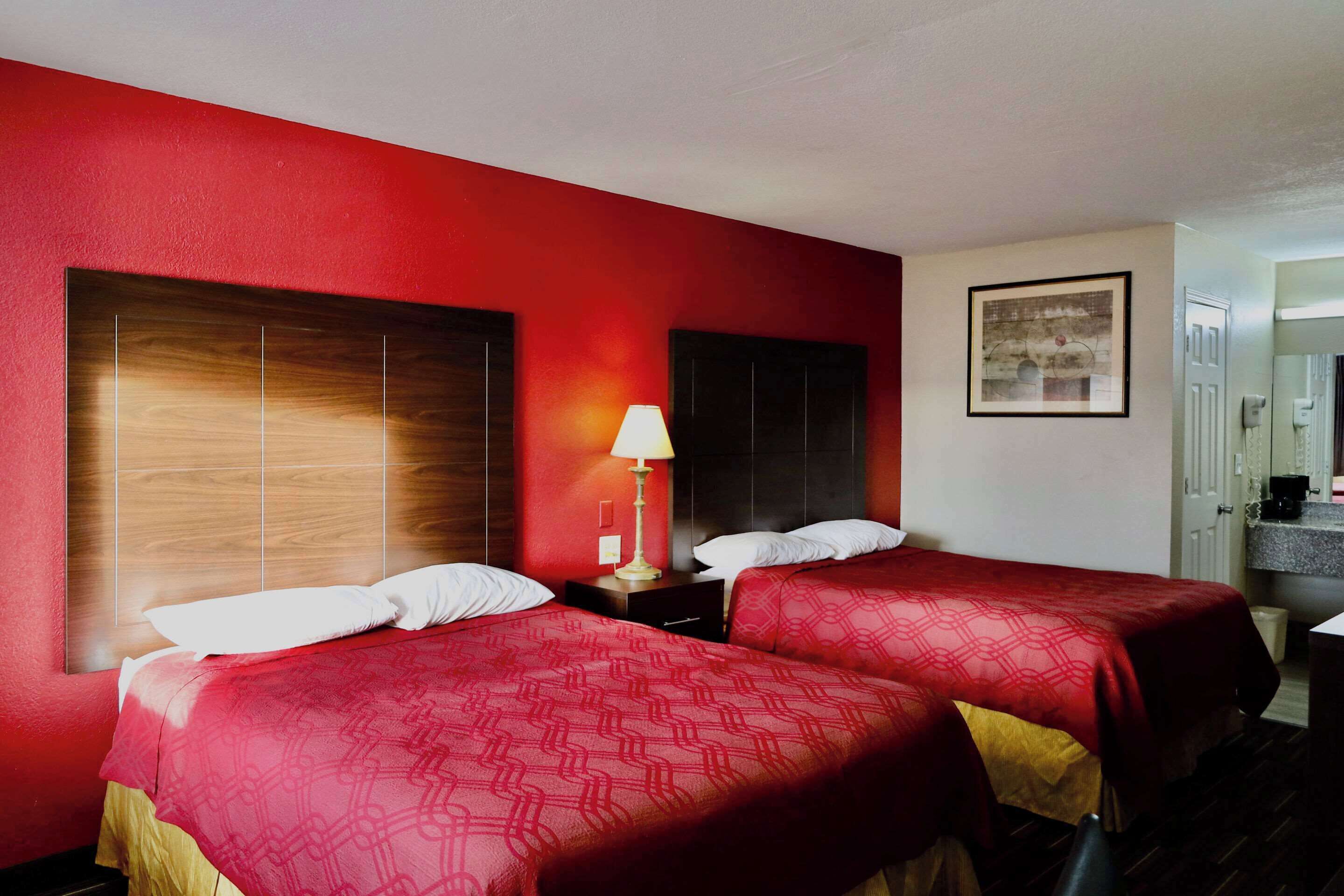A hotel room with two beds, a nightstand with a lamp, a framed picture on the wall, and a bathroom visible in the background.