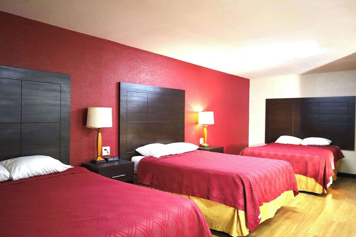 Une chambre d’hôtel avec trois lits, chacun recouvert d’une couverture rouge et agrémenté d’oreillers blancs. Il y a deux tables de chevet avec des lampes. Les murs sont peints en rouge et le plafond est blanc.