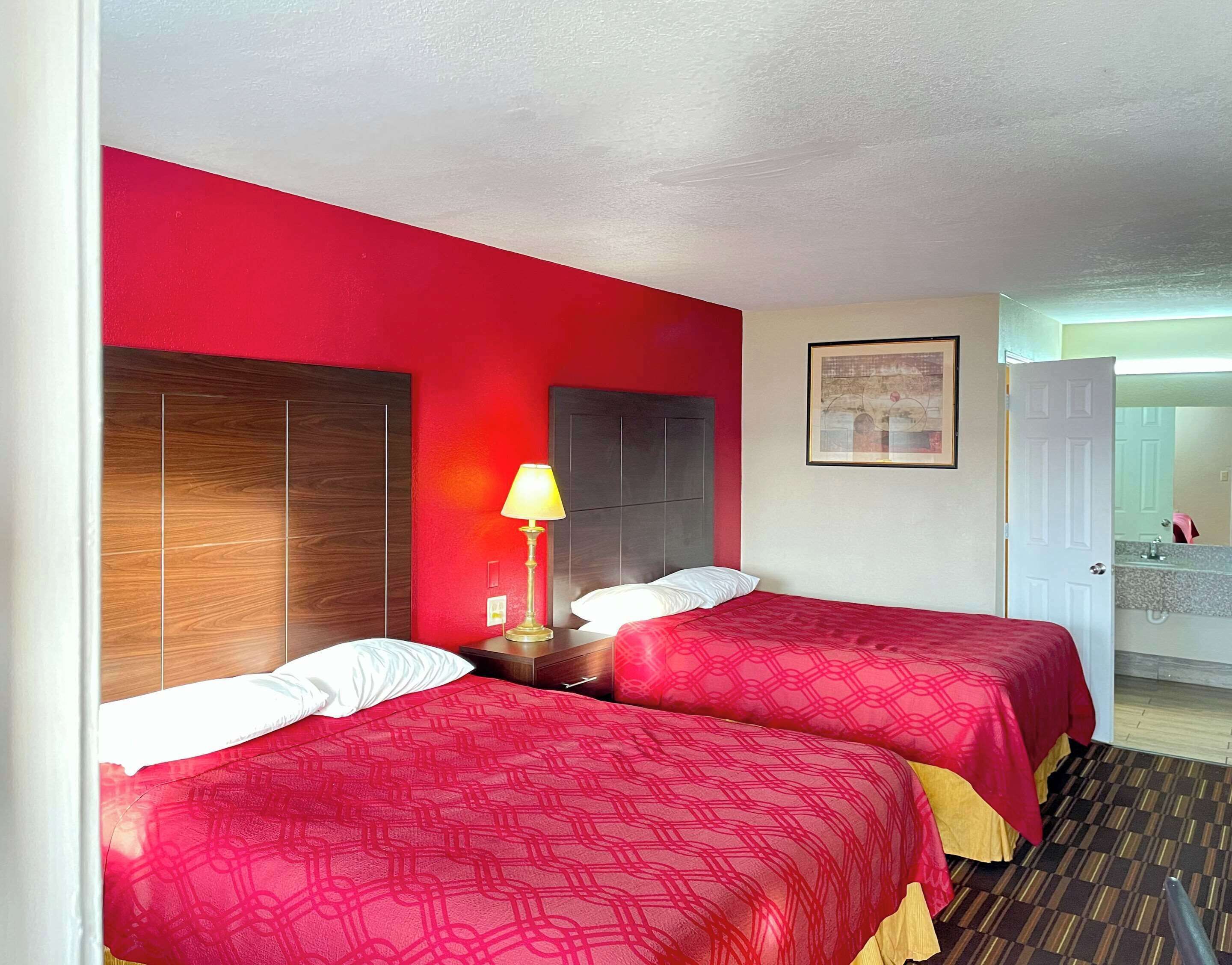 A hotel room with two beds, a nightstand with a lamp, a bathroom visible through an open door, and a framed picture on the wall.