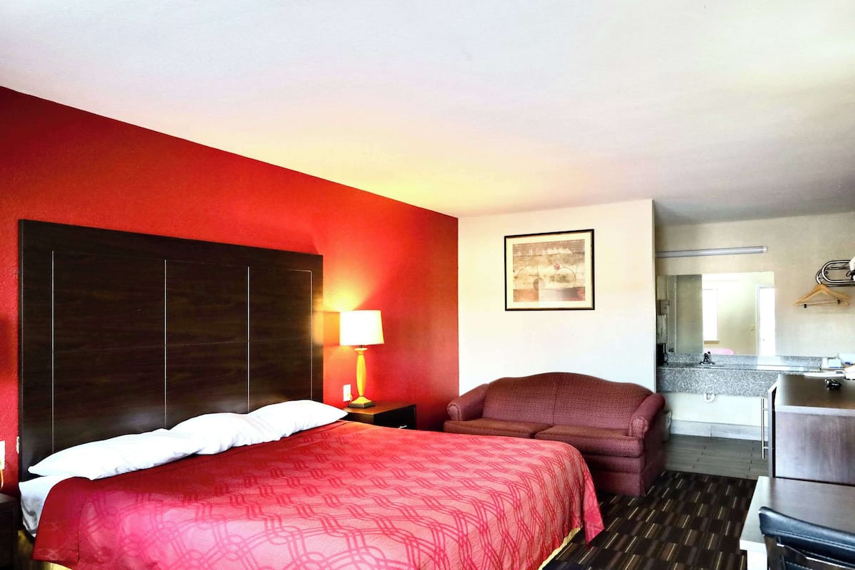 Une chambre d’hôtel avec un grand lit, un bureau, une chaise, une table de chevet avec une lampe, et une salle de bain visible à travers une porte ouverte.