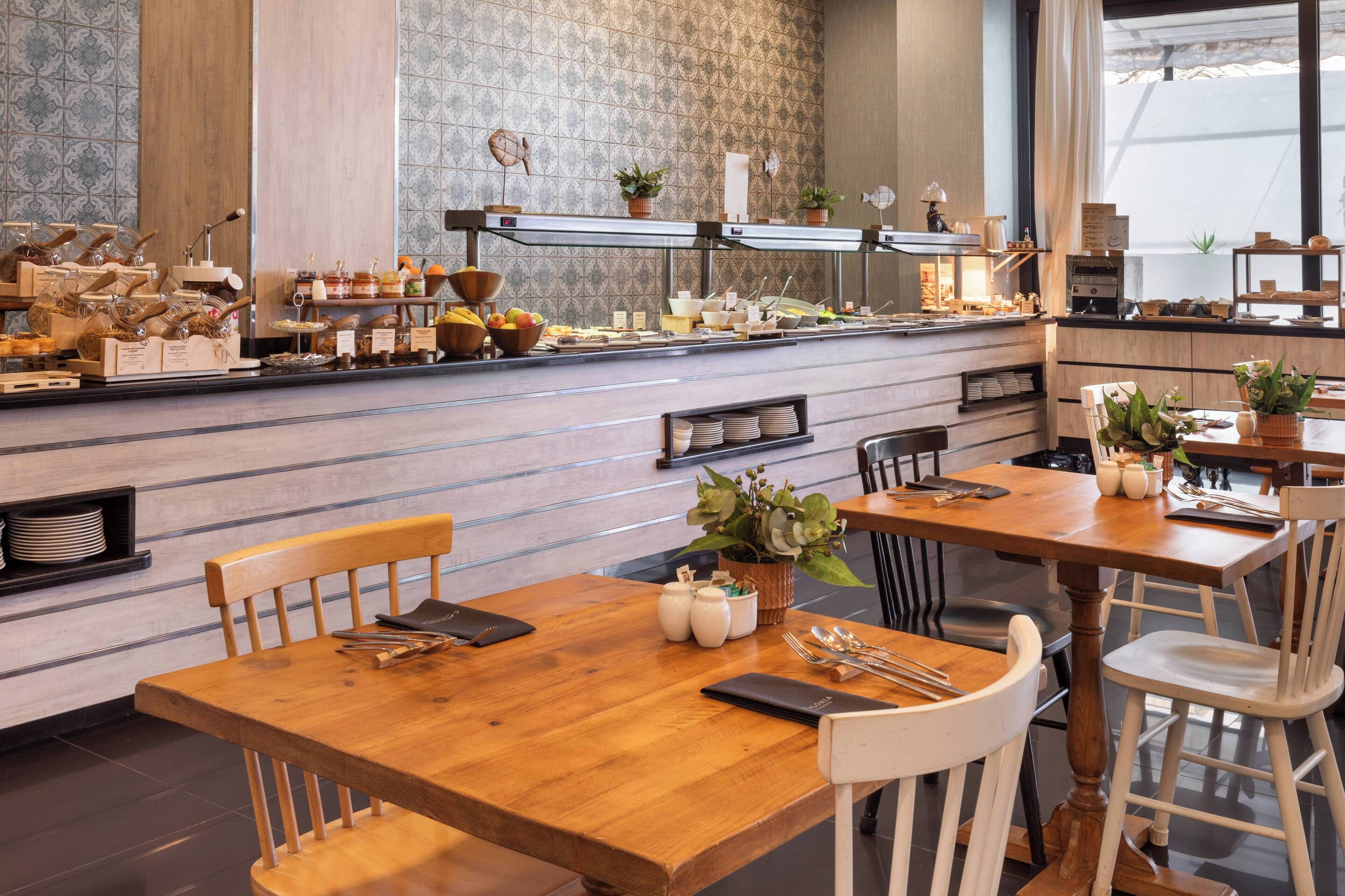 Un restaurante con mesas y sillas de madera, una barra de buffet con diferentes platillos, y un muro con diseño.