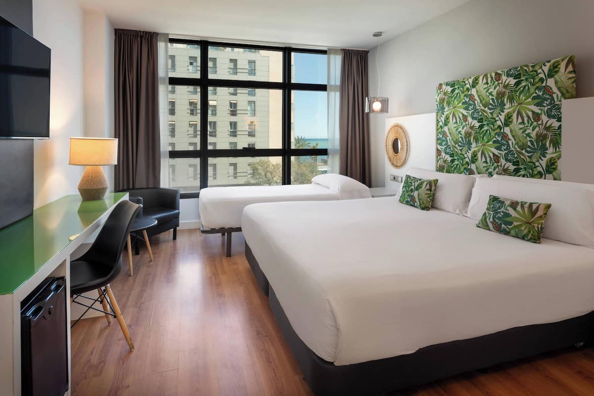 A hotel room with two beds, a desk with a lamp, a chair, and a large window with a view of buildings and the sea.