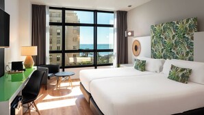Habitació d'hotel amb un llit gran, escriptori amb cadira, una taula petita i vista a l'oceà a través de grans finestres.