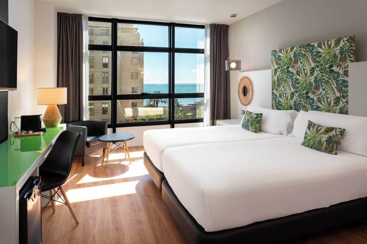 A hotel room with two beds, a desk, a chair, a small table, and a large window offering a view of the ocean.