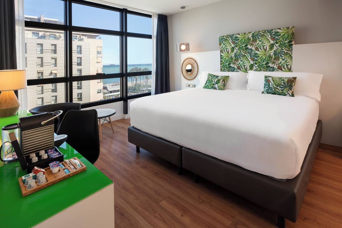 A hotel room with a large bed, a desk with a coffee machine, and a view of the city through large windows.