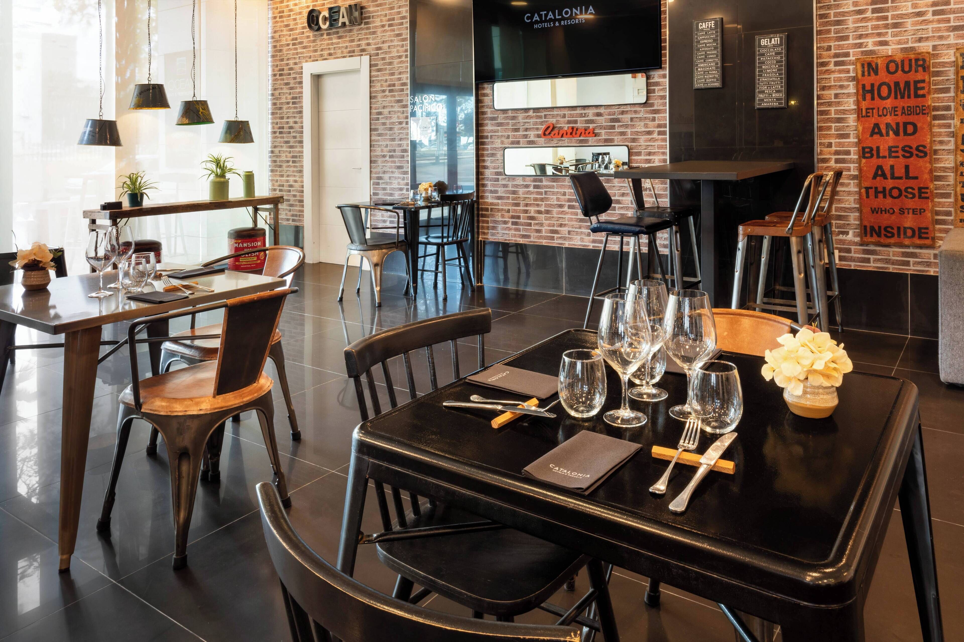 Un espace repas moderne avec des tables et des chaises noires, un mur en briques et une enseigne indiquant « CATALONIA HOTELS & RESORTS ».
